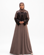 A woman modeling a long taupe-brown abaya dress with black lace sleeves and a matching taupe hijab, standing against a plain white studio background.