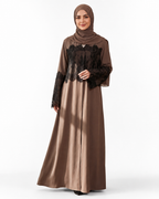 A woman modeling a long-sleeved brown abaya dress with black lace overlay on the bodice and sleeves, paired with a matching brown hijab, against a plain white studio background.