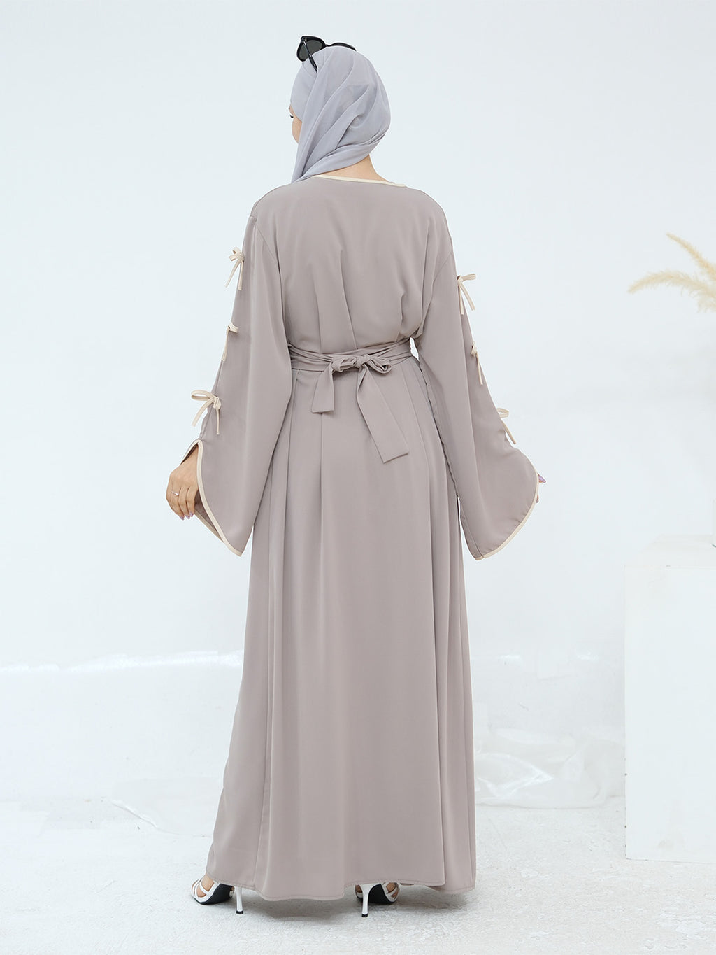 A woman viewed from the back wearing a long taupe-gray abaya dress with wide sleeves, tie details at the cuffs, and a matching waist tie, paired with a light gray hijab.