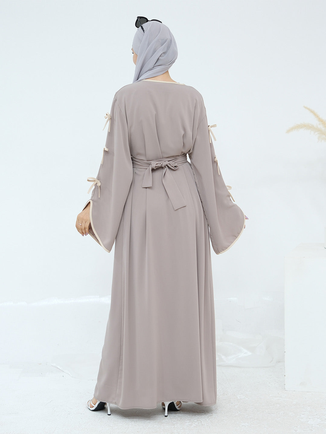 A woman viewed from the back wearing a long taupe-gray abaya dress with wide sleeves, tie details at the cuffs, and a matching waist tie, paired with a light gray hijab.