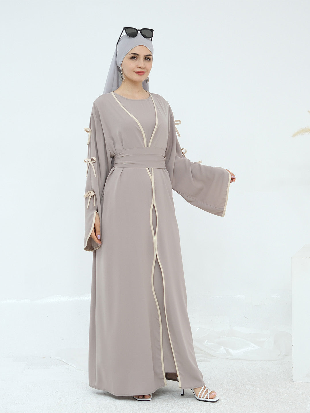 A woman modeling a long taupe-gray abaya dress with cream-colored trim and tie details on the sleeves, paired with a matching headscarf and sunglasses.