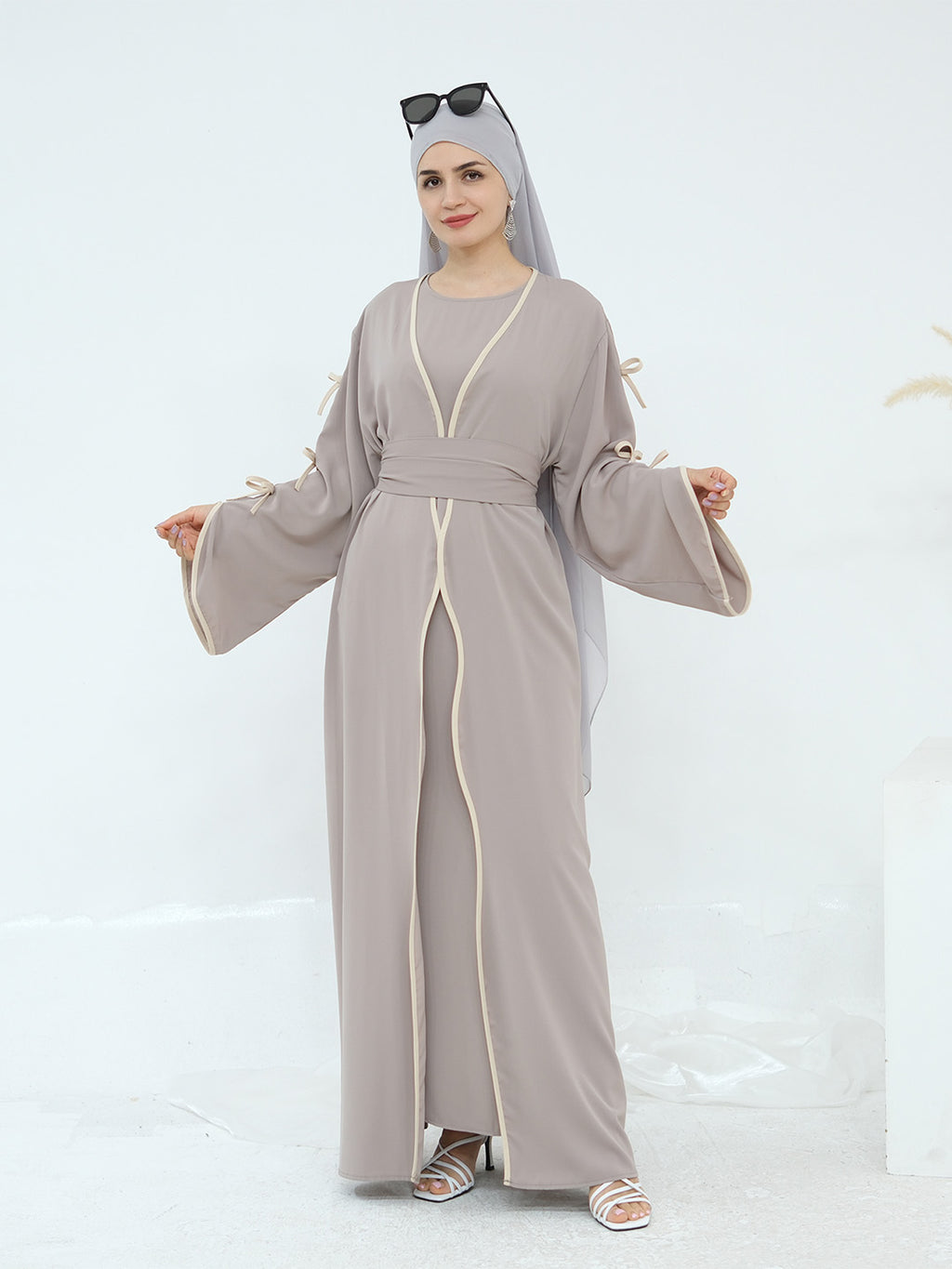 A woman modeling a taupe-gray abaya with cream-colored trim and a matching headscarf, standing against a clean white studio background.
