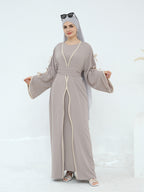 A woman modeling a taupe-gray abaya with cream-colored trim and a matching headscarf, standing against a clean white studio background.