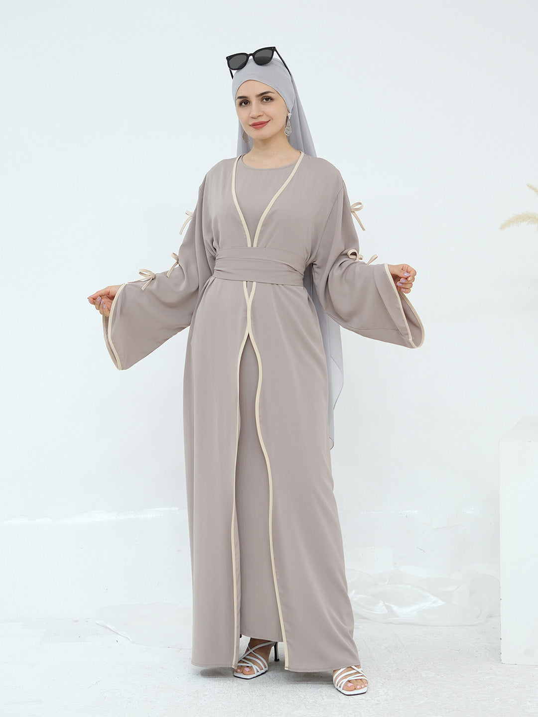 A woman modeling a taupe-gray abaya with cream-colored trim and a matching headscarf, standing against a clean white studio background.