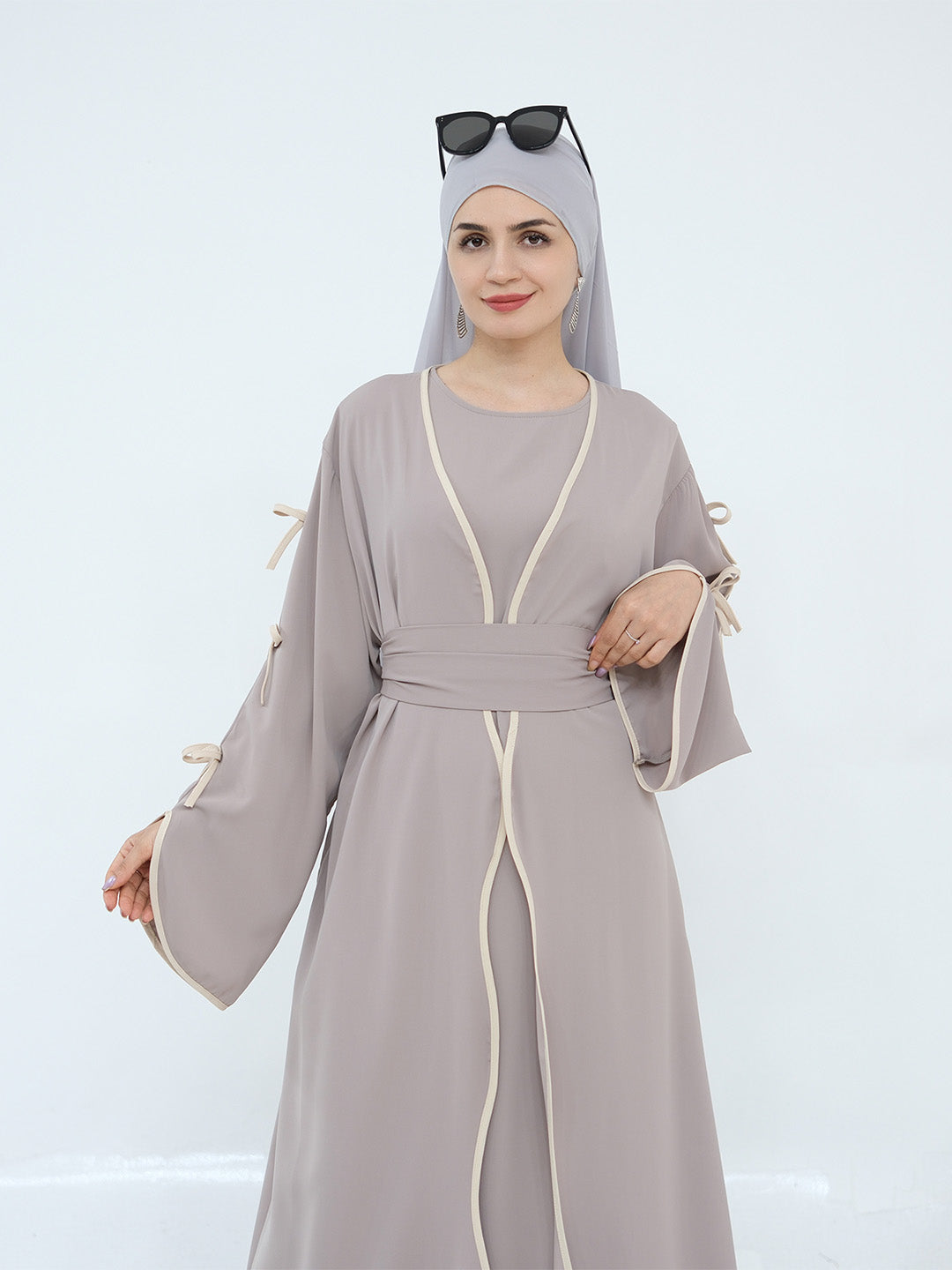 A woman modeling a light gray abaya dress with cream-colored piping details, a matching gray hijab, and black sunglasses on her head, set against a plain white studio background.