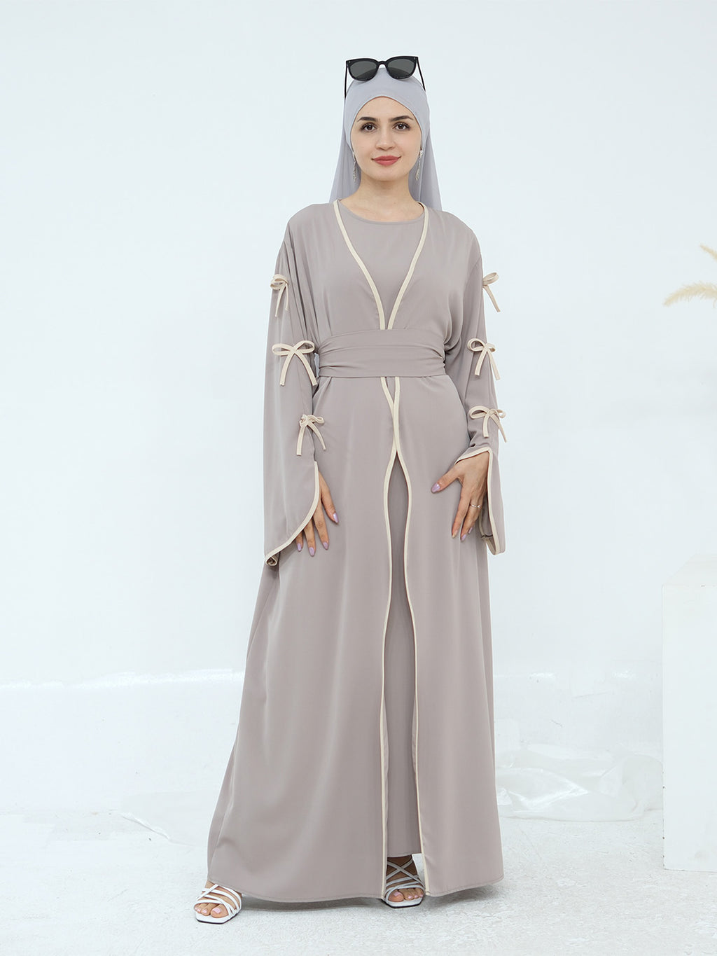 A woman modeling a long taupe-gray abaya dress with cream-colored trim and tie details on the sleeves, paired with a matching hijab and sunglasses.