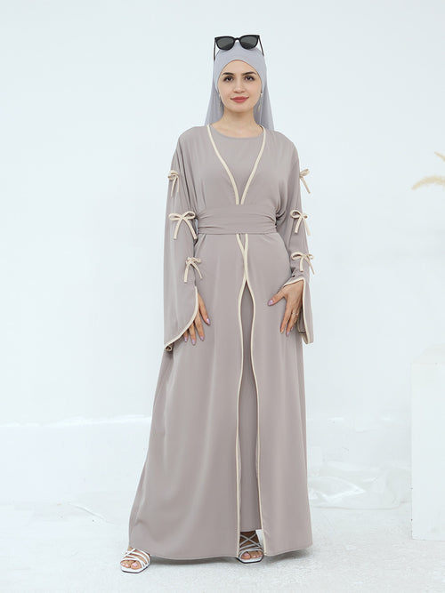 A woman modeling a long taupe-gray abaya dress with cream-colored trim and tie details on the sleeves, paired with a matching hijab and sunglasses.