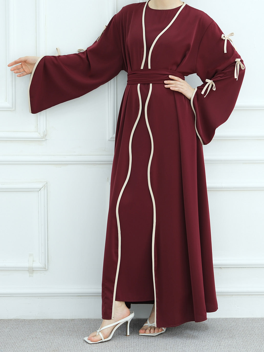 A woman modeling a long maroon abaya dress with cream-colored wavy piping details and matching tie straps on the sleeves, paired with white heeled sandals against a white paneled wall.