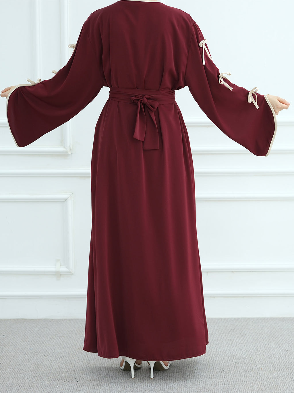A woman modeling a long, deep burgundy maxi dress with wide bell sleeves, cream tie details on the sleeves, and a matching waist tie, shown from the back against a white paneled wall.