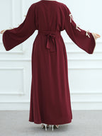 A woman modeling a long, deep burgundy maxi dress with wide bell sleeves, cream tie details on the sleeves, and a matching waist tie, shown from the back against a white paneled wall.