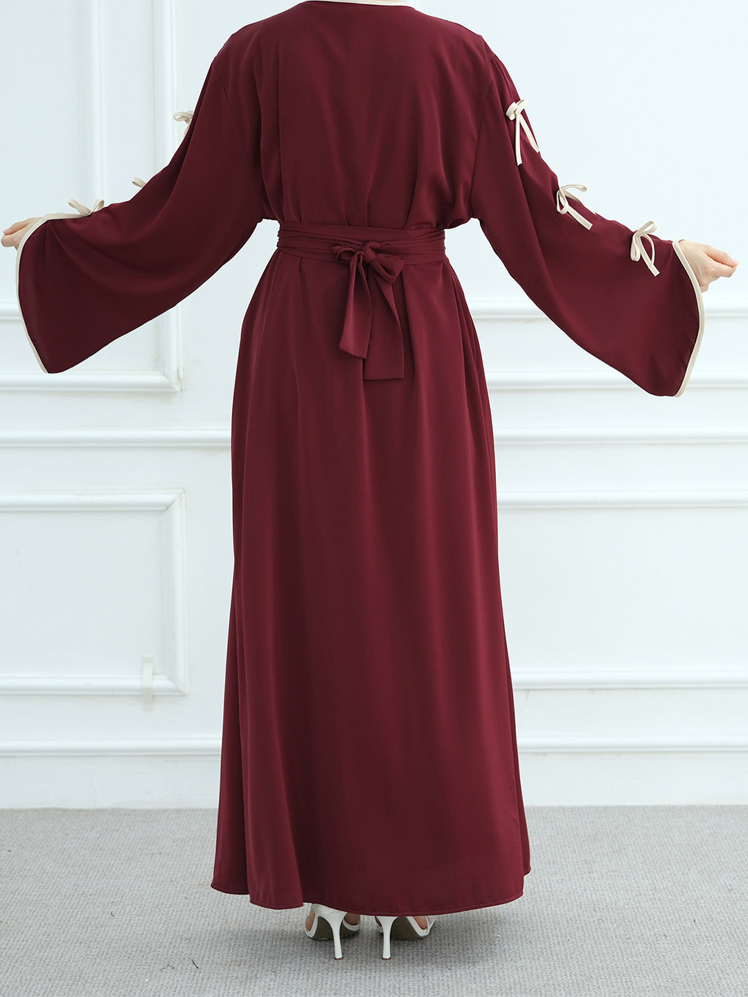 A woman modeling a long, deep burgundy maxi dress with wide bell sleeves, cream tie details on the sleeves, and a matching waist tie, shown from the back against a white paneled wall.