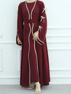A woman wearing a long maroon maxi dress with cream-colored piping, tie details on the sleeves, and a matching waist belt, standing against a white paneled wall.