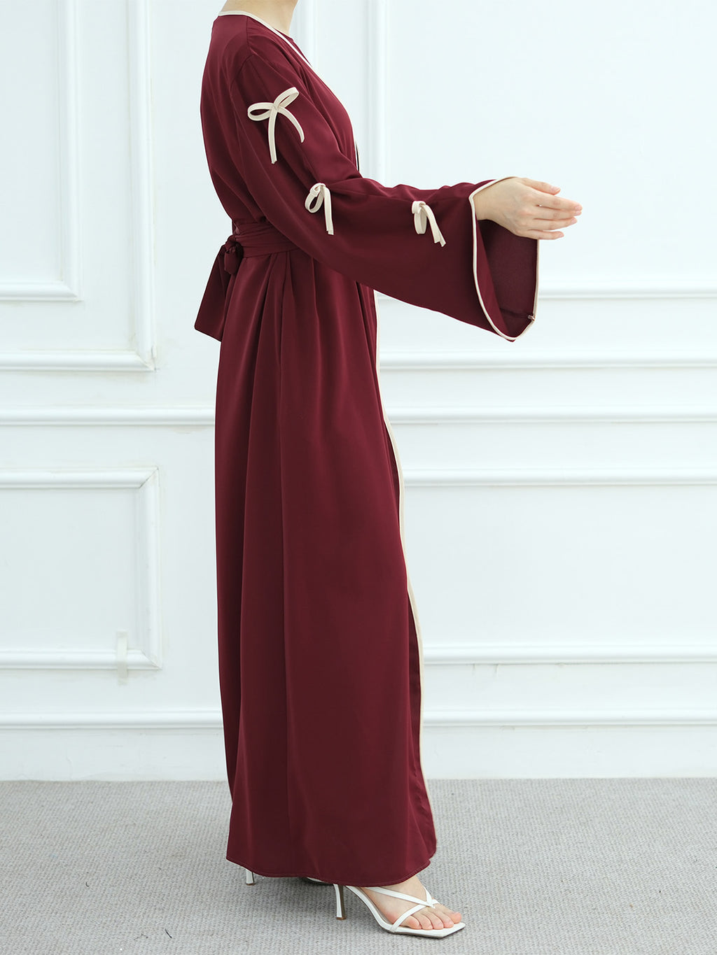 A woman wearing a long-sleeved maroon maxi dress with cream-colored tie details on the sleeves and a matching waist tie, standing against a white paneled wall.