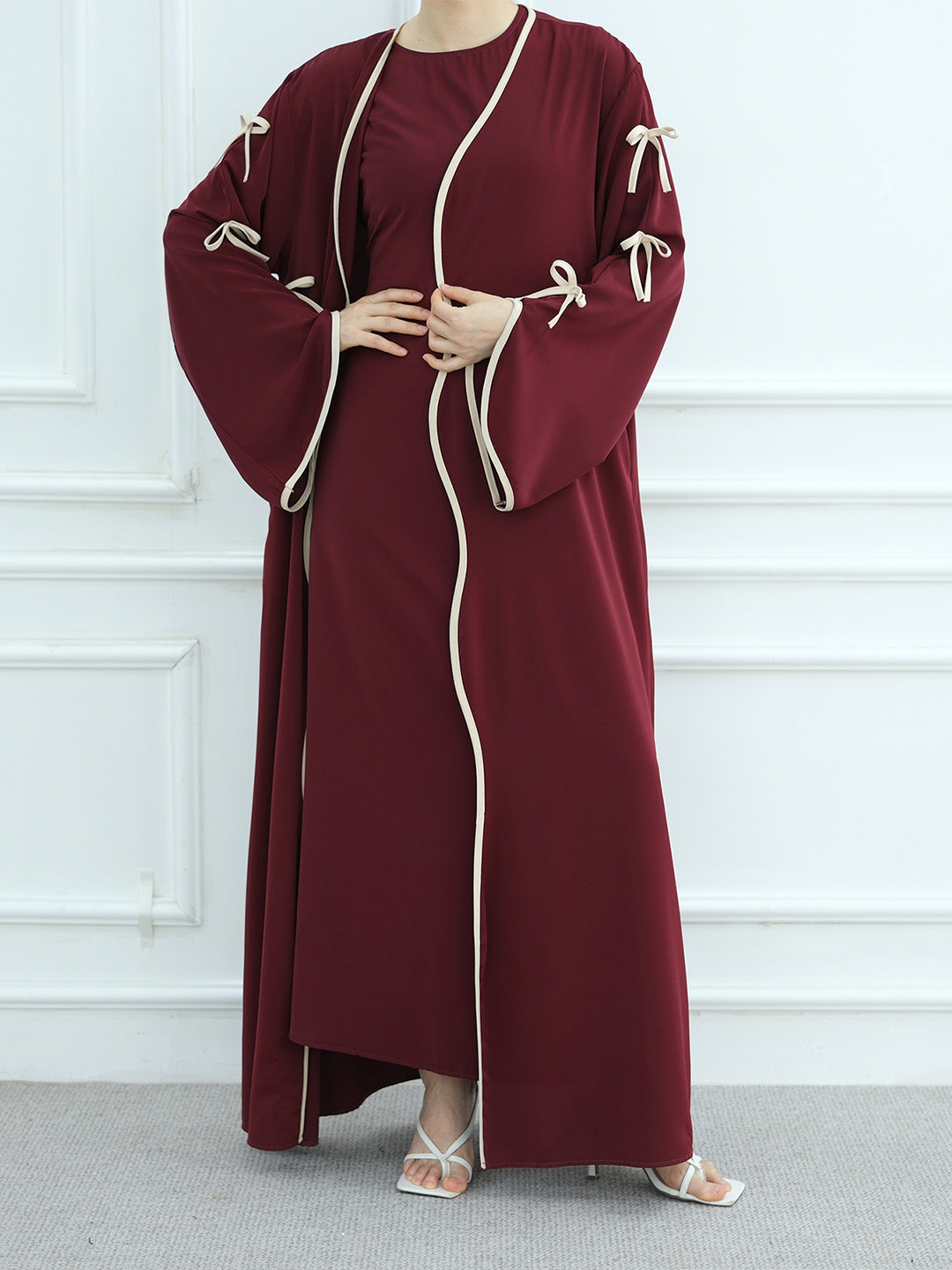 A woman wearing a long burgundy abaya dress with cream-colored piping and tie details on the sleeves, standing against a white paneled wall.