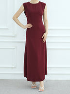 A woman modeling a sleeveless, floor-length maroon maxi dress against a white paneled wall background.