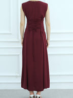 A sleeveless, floor-length burgundy dress shown from the back on a model, featuring a lace-up waist and tie detail, set against a white paneled wall.