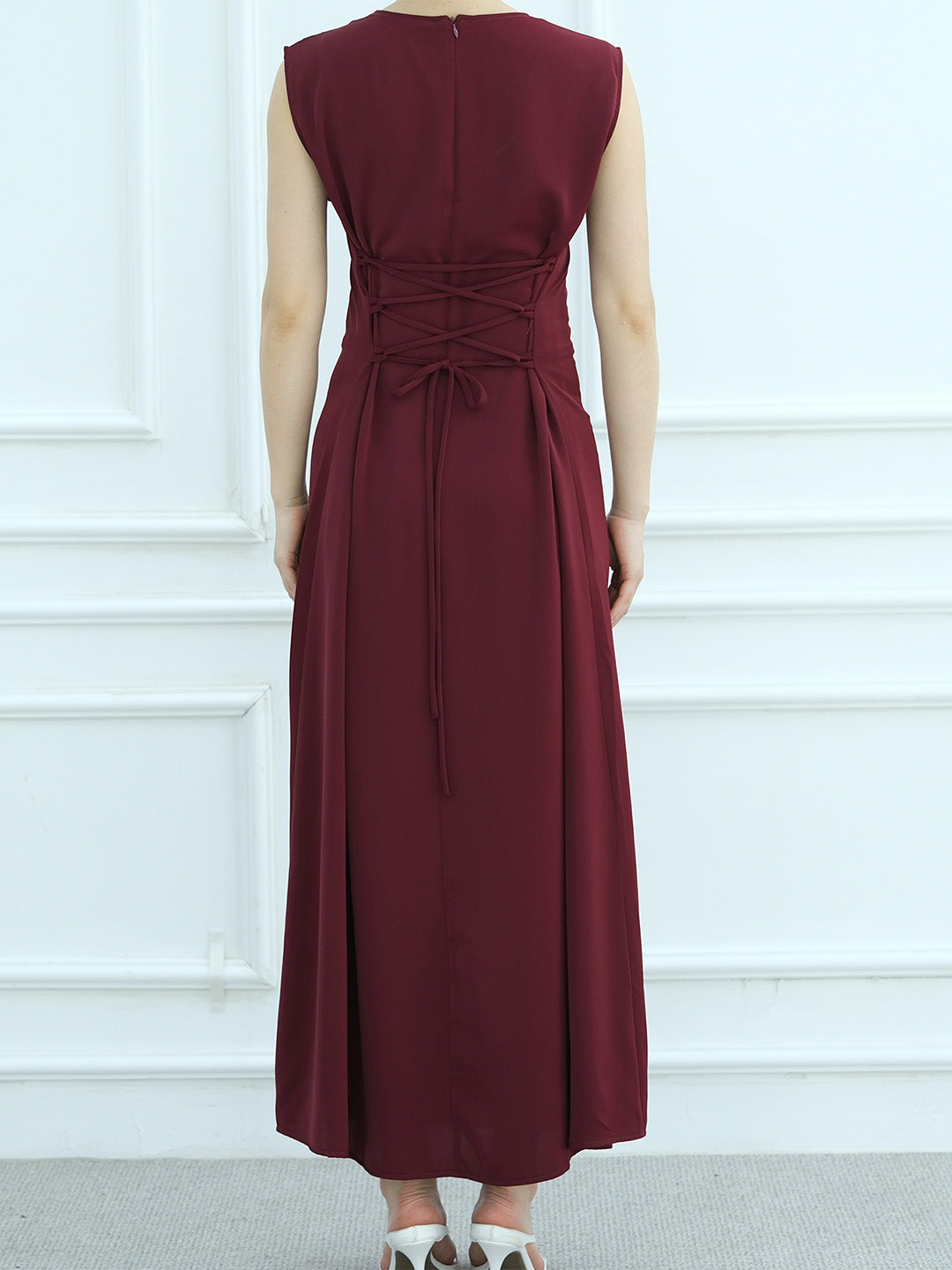 A sleeveless, floor-length burgundy dress shown from the back on a model, featuring a lace-up waist and tie detail, set against a white paneled wall.