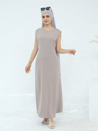 A woman modeling a sleeveless, floor-length taupe maxi dress with a matching headscarf and sunglasses on her head, paired with strappy sandals, set against a clean white studio background.