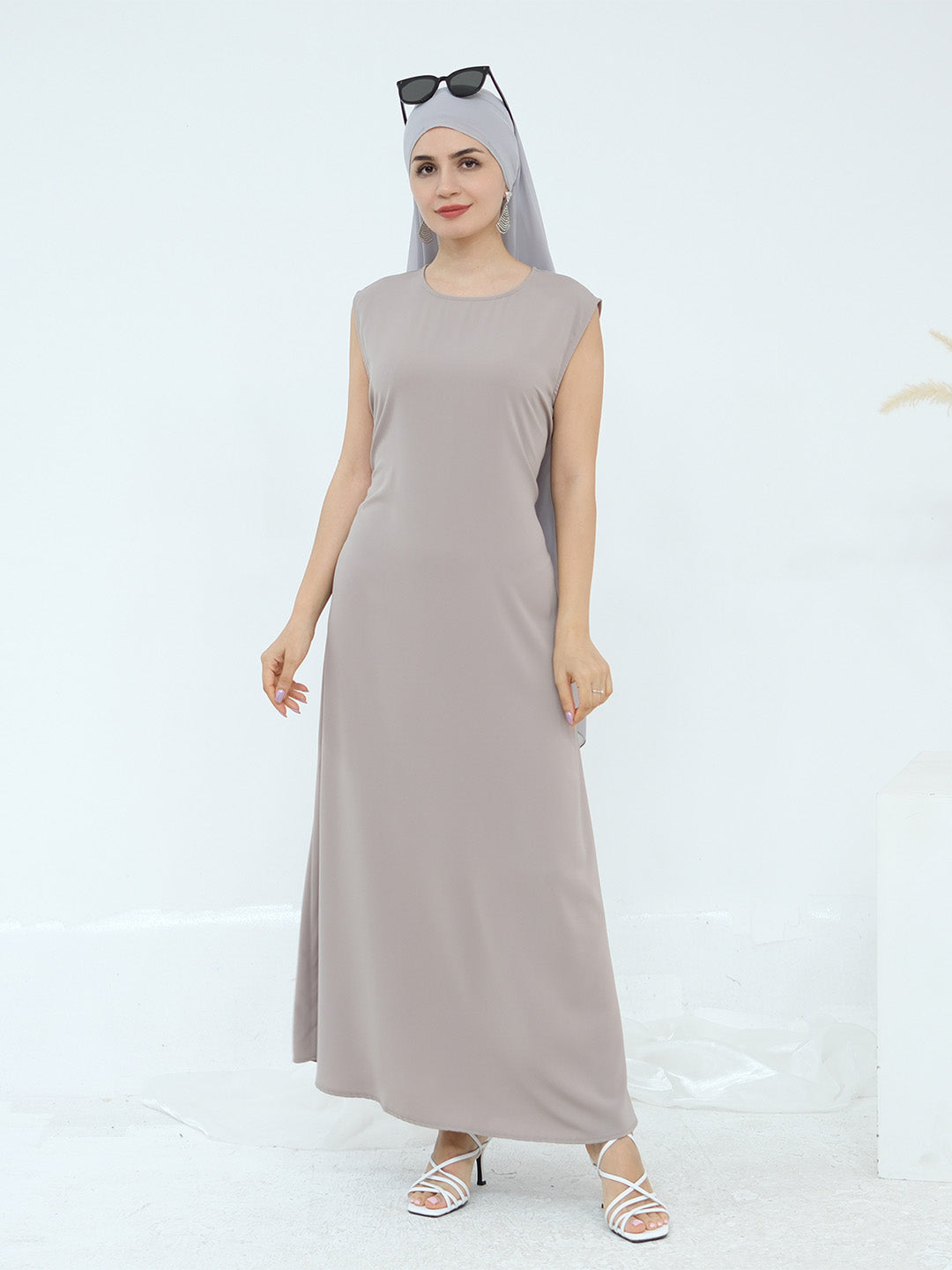 A woman modeling a sleeveless, floor-length taupe maxi dress with a matching hijab and white strappy heels against a clean white studio background.