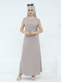 A woman modeling a sleeveless, floor-length taupe maxi dress with a matching hijab and sunglasses on her head, paired with white strappy heels against a clean white studio background.