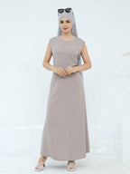 A woman modeling a sleeveless, floor-length taupe maxi dress with a matching hijab and sunglasses on her head, paired with white strappy heels against a clean white studio background.