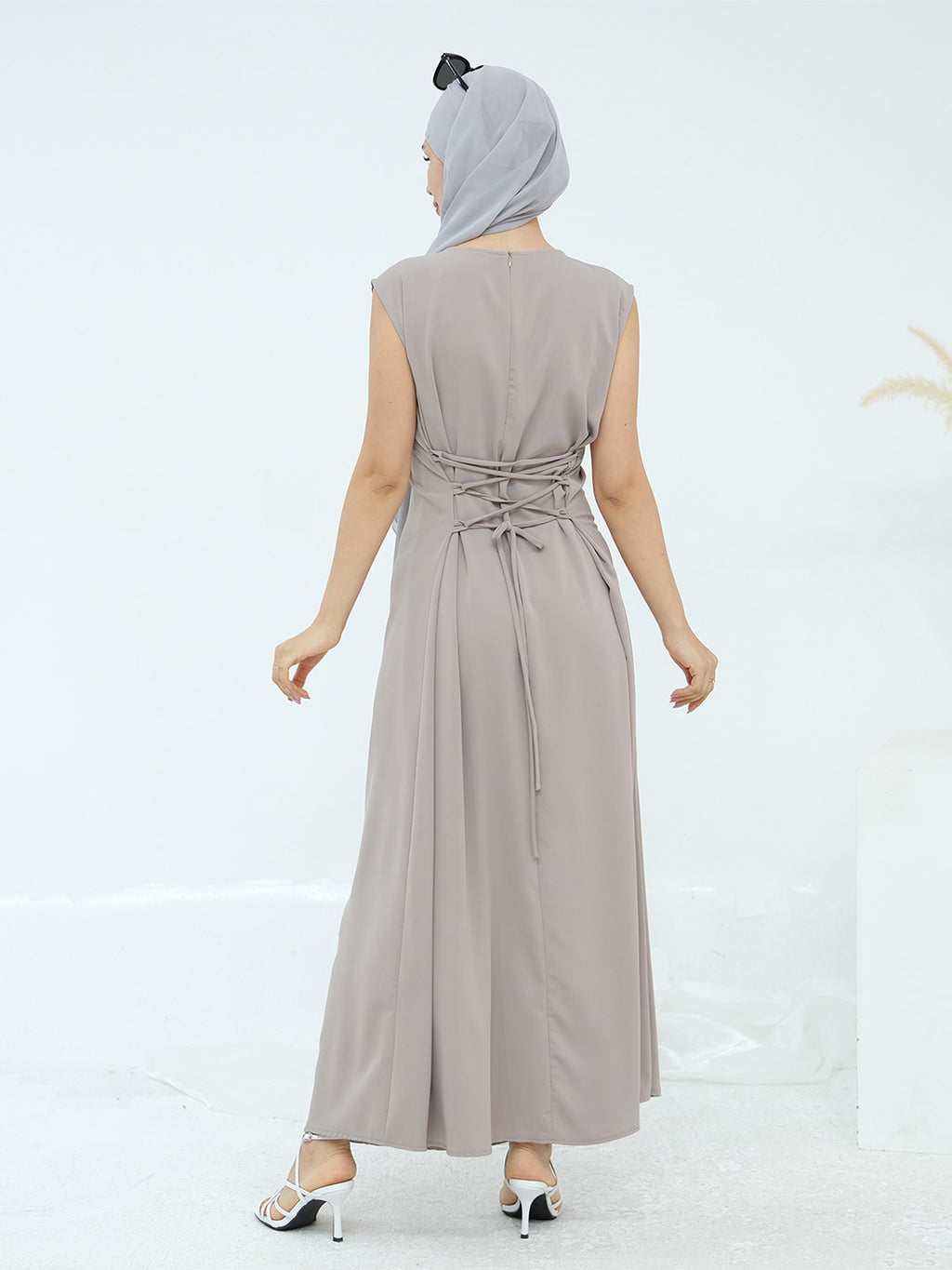 A woman shown from the back wearing a sleeveless, floor-length taupe maxi dress with a matching headscarf and white heeled sandals, set against a clean white studio background.