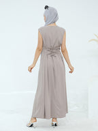 A woman shown from the back wearing a sleeveless, floor-length taupe maxi dress with a matching headscarf and white heeled sandals, set against a clean white studio background.