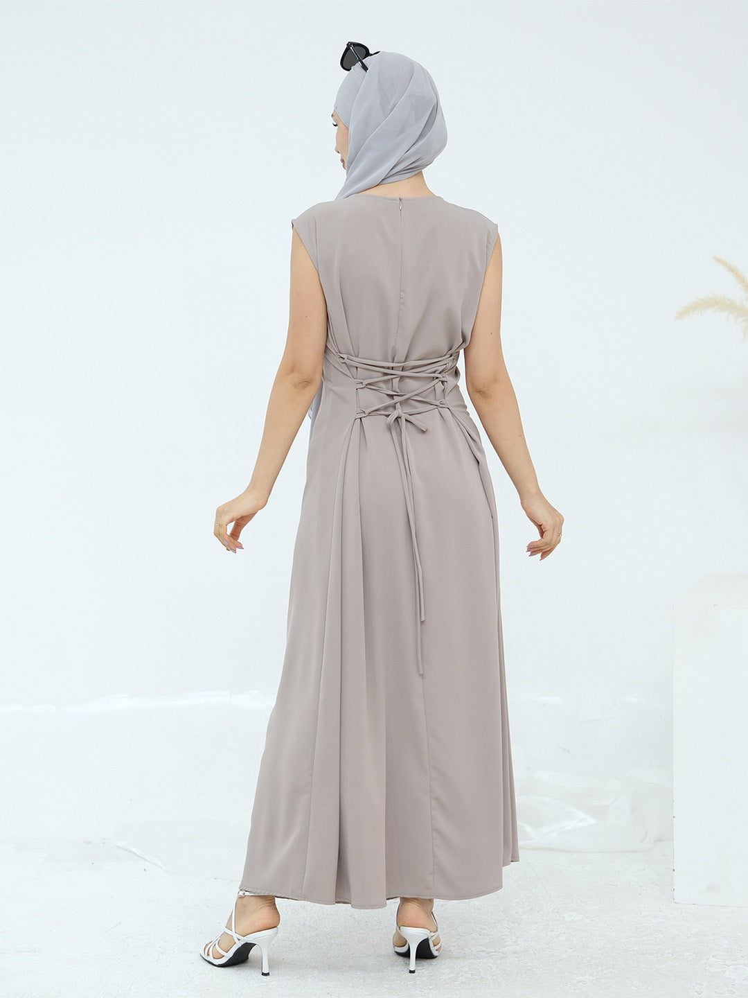 A woman shown from the back wearing a sleeveless, floor-length taupe maxi dress with a matching headscarf and white heeled sandals, set against a clean white studio background.